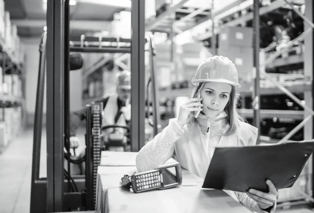 Female warehouse staff wearing hard hat speaking on phone at workstation